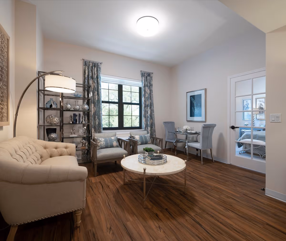 A cozy living room in a senior living facility featuring a beige tufted sofa, two cushioned armchairs with patterned pillows, a round marble coffee table with decorative items, a tall floor lamp, and a metal shelving unit with various decorative objects. The room has wood flooring, a large window with floral curtains, and a small dining area with a glass table and two upholstered chairs. A glass-paneled door leads to a bedroom visible in the background.