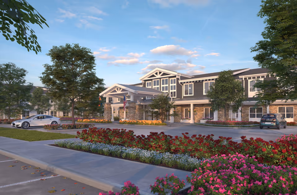 Front exterior of a two-story senior living building with a porte-cochère, landscaped flowerbeds, trees, and cars in the driveway.