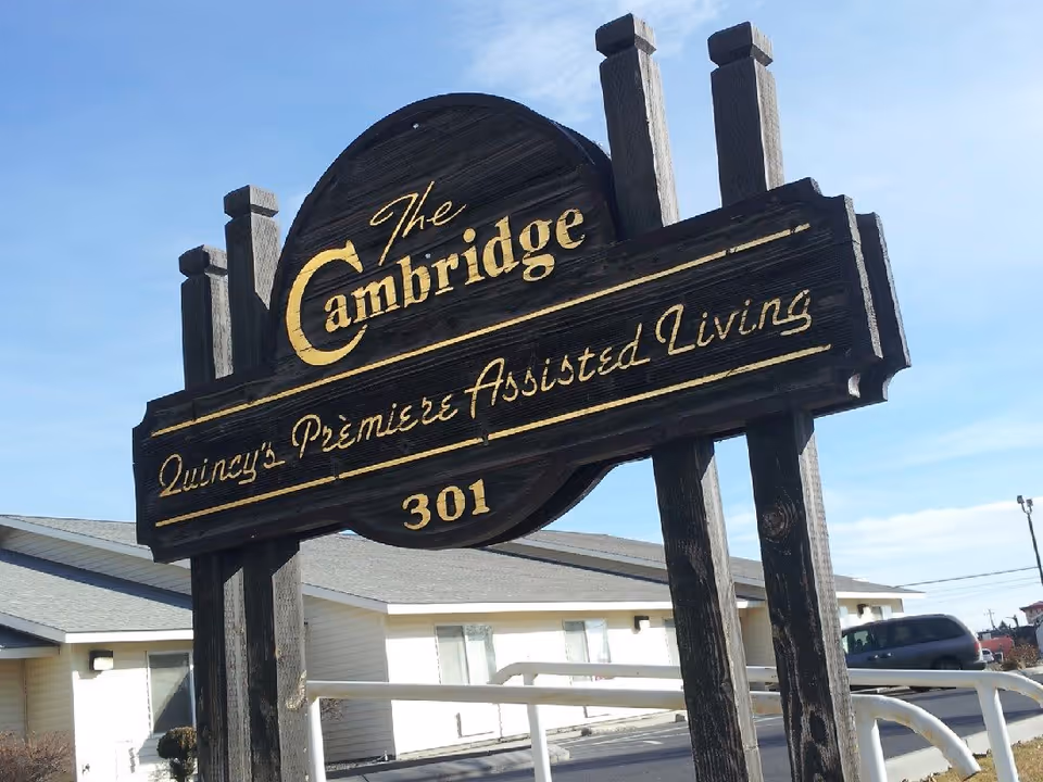 A wooden sign for The Cambridge Assisted Living, labeled as Quincy’s Premiere Assisted Living, with the number 301. The sign is mounted on wooden posts and is set against a background of a single-story building and a clear blue sky.
