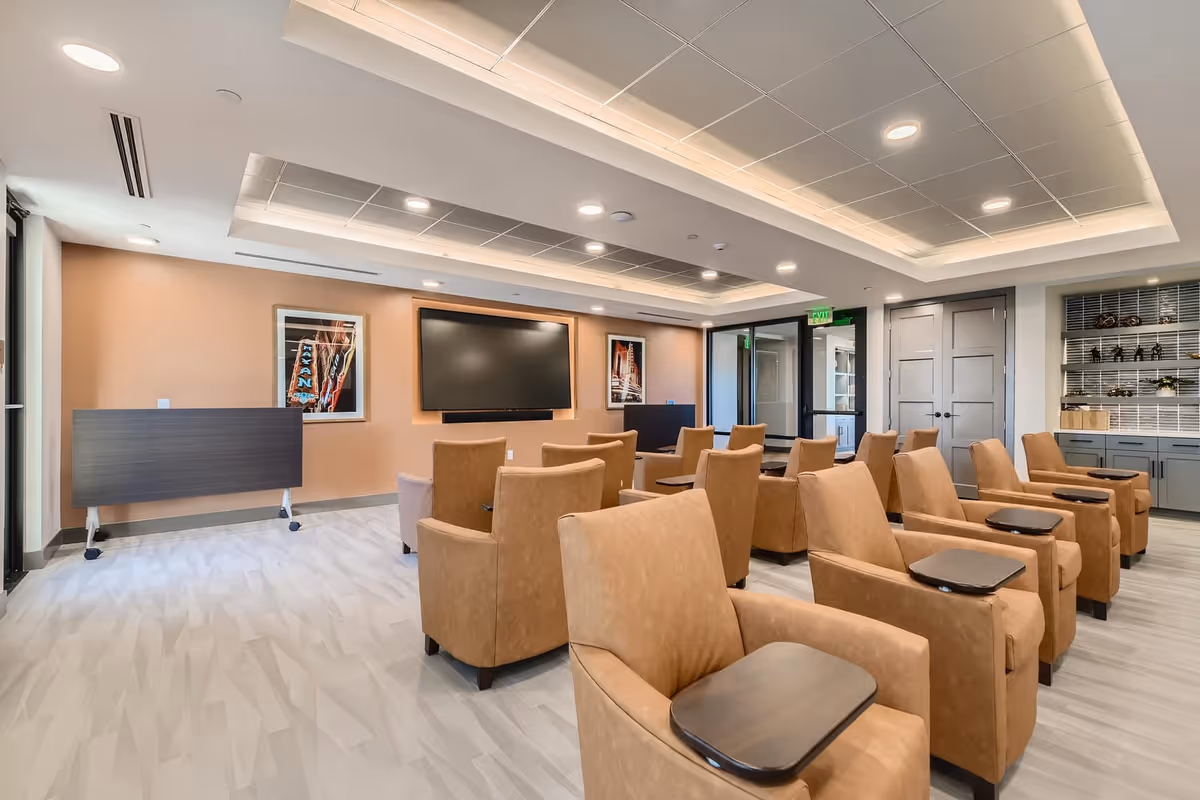 A modern senior living media room with rows of tan recliner chairs facing a large wall-mounted TV and soft recessed lighting.