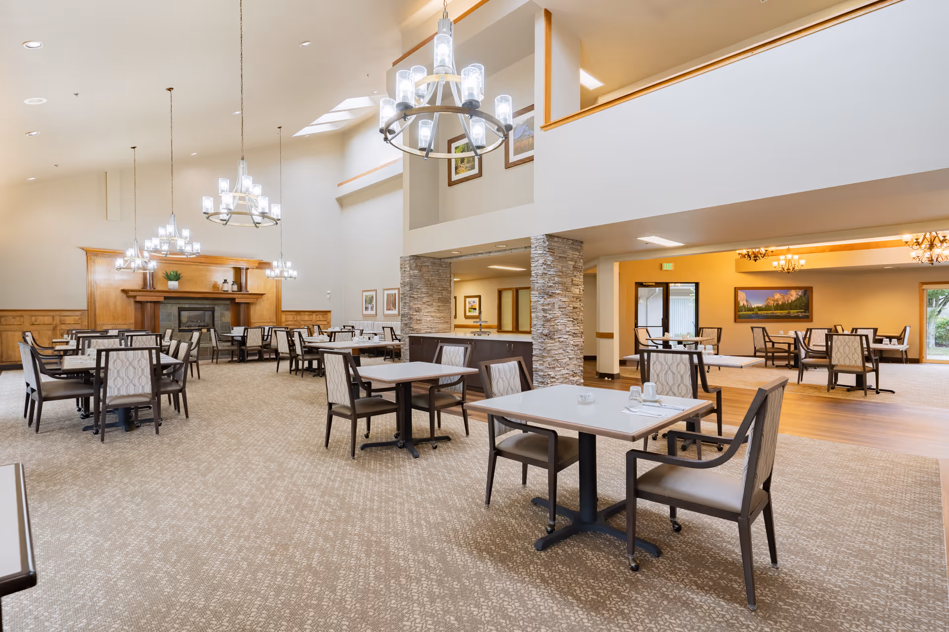 A spacious dining room with multiple tables and chairs arranged neatly. The room features high ceilings with modern chandeliers, a fireplace with a wooden mantel, and stone pillars. The walls are decorated with framed pictures, and there are large windows allowing natural light to enter.