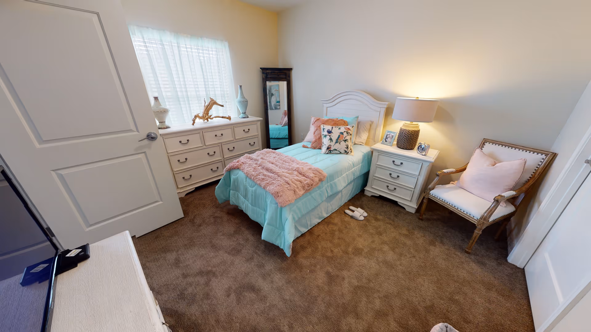 A cozy bedroom with a single bed dressed in light blue bedding and a pink throw blanket. The room features a white dresser with decorative items, a full-length mirror, a nightstand with a lamp and framed photos, a cushioned wooden chair with a pink pillow, and brown carpeted flooring. Natural light filters through a window with sheer white curtains.