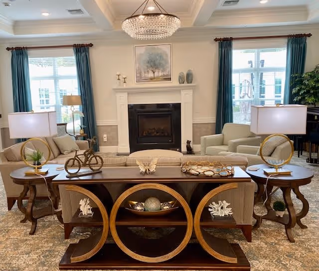 A cozy living room with a central fireplace, flanked by two large windows with teal curtains. The room features a beige sofa and armchairs, two side tables with lamps, and a decorative wooden console table with circular designs and various ornaments. A chandelier hangs from the ceiling, and a framed artwork is above the fireplace.