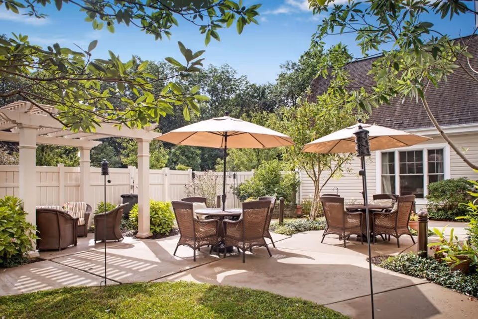 Outdoor patio area with two round tables each surrounded by four wicker chairs and shaded by large beige umbrellas. There is a white pergola with two cushioned armchairs underneath, surrounded by green plants and trees. A white fence encloses the space, and a building with white siding and windows is visible in the background under a clear blue sky.