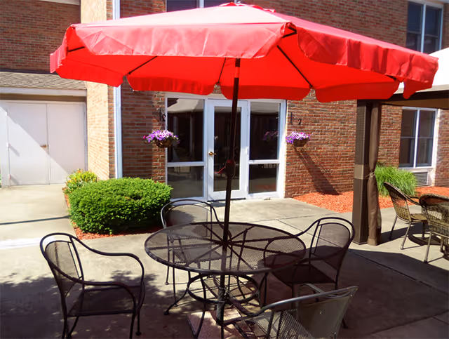 Outdoor patio area with a round metal table and four metal chairs under a large red umbrella. The patio is adjacent to a brick building with glass doors and windows. There are hanging flower baskets and some green bushes near the building.