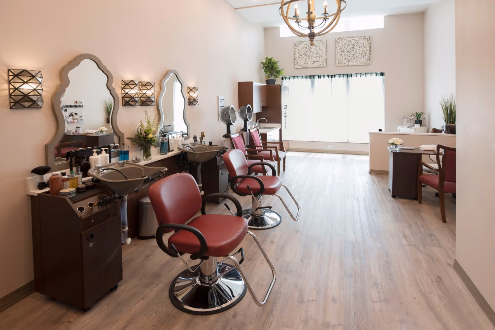 Interior view of a salon area with three red salon chairs in front of wash basins and large decorative mirrors on the wall. The room has wooden flooring, soft lighting fixtures, and a large window with sheer curtains letting in natural light. There are plants and decorative wall art adding to the cozy atmosphere.