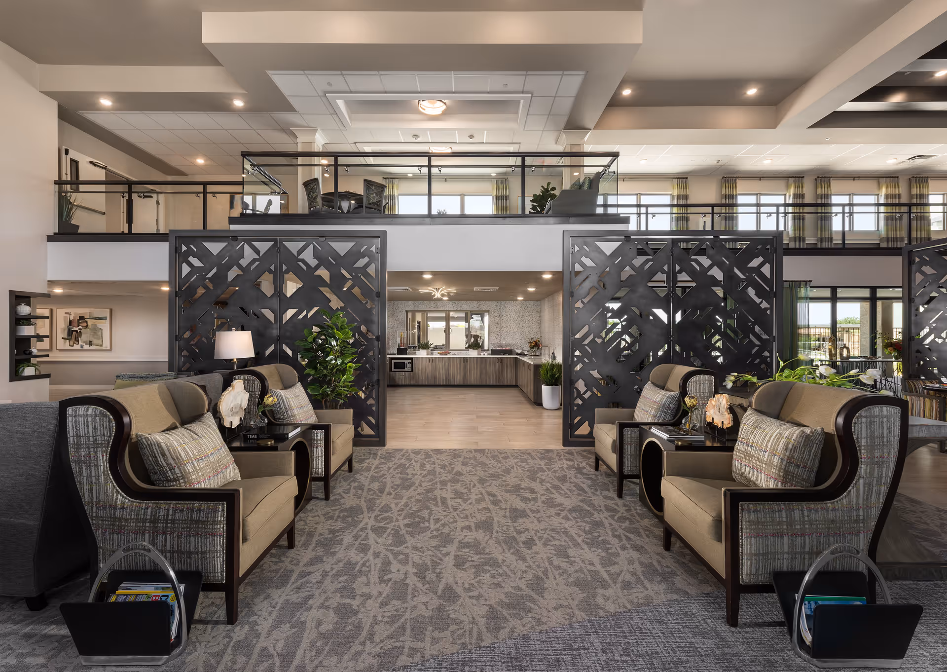 Spacious and modern senior living facility common area with comfortable armchairs arranged in pairs on either side of a patterned carpet. Decorative black partition screens separate the seating area from a kitchen or dining space in the background. The room features high ceilings with recessed lighting, large windows with curtains, and an upper balcony area with additional seating and plants.