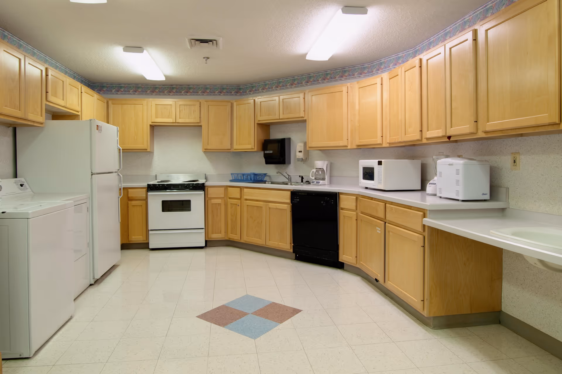 A spacious kitchen with light wood cabinets, white appliances including a refrigerator, stove, microwave, dishwasher, and washing machine. The floor has a tiled pattern with a diamond shape in the center. The room is well-lit with fluorescent ceiling lights.