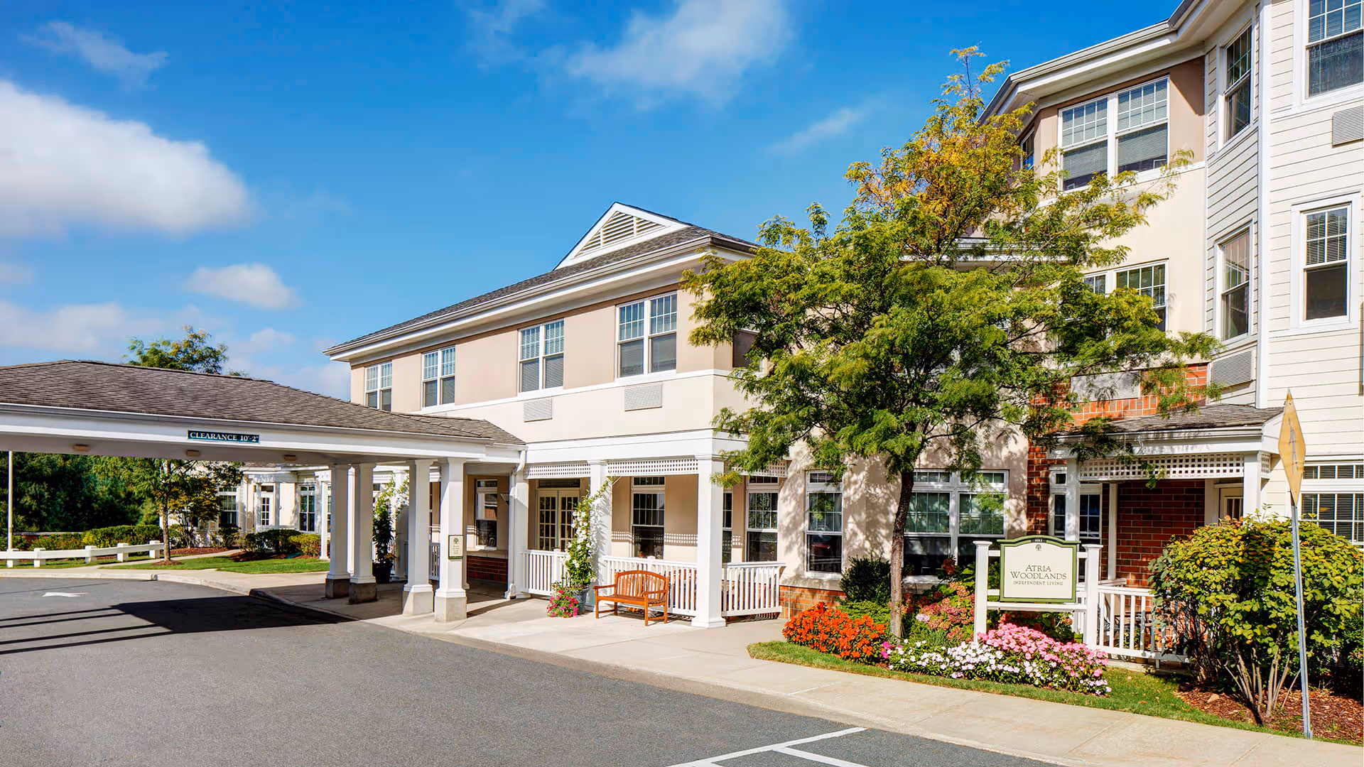 Exterior view of Atria Woodlands senior living facility showing a two-story building with beige and white siding, a covered entrance with columns, a bench, and landscaped flower beds with a tree and colorful flowers under a blue sky with some clouds.