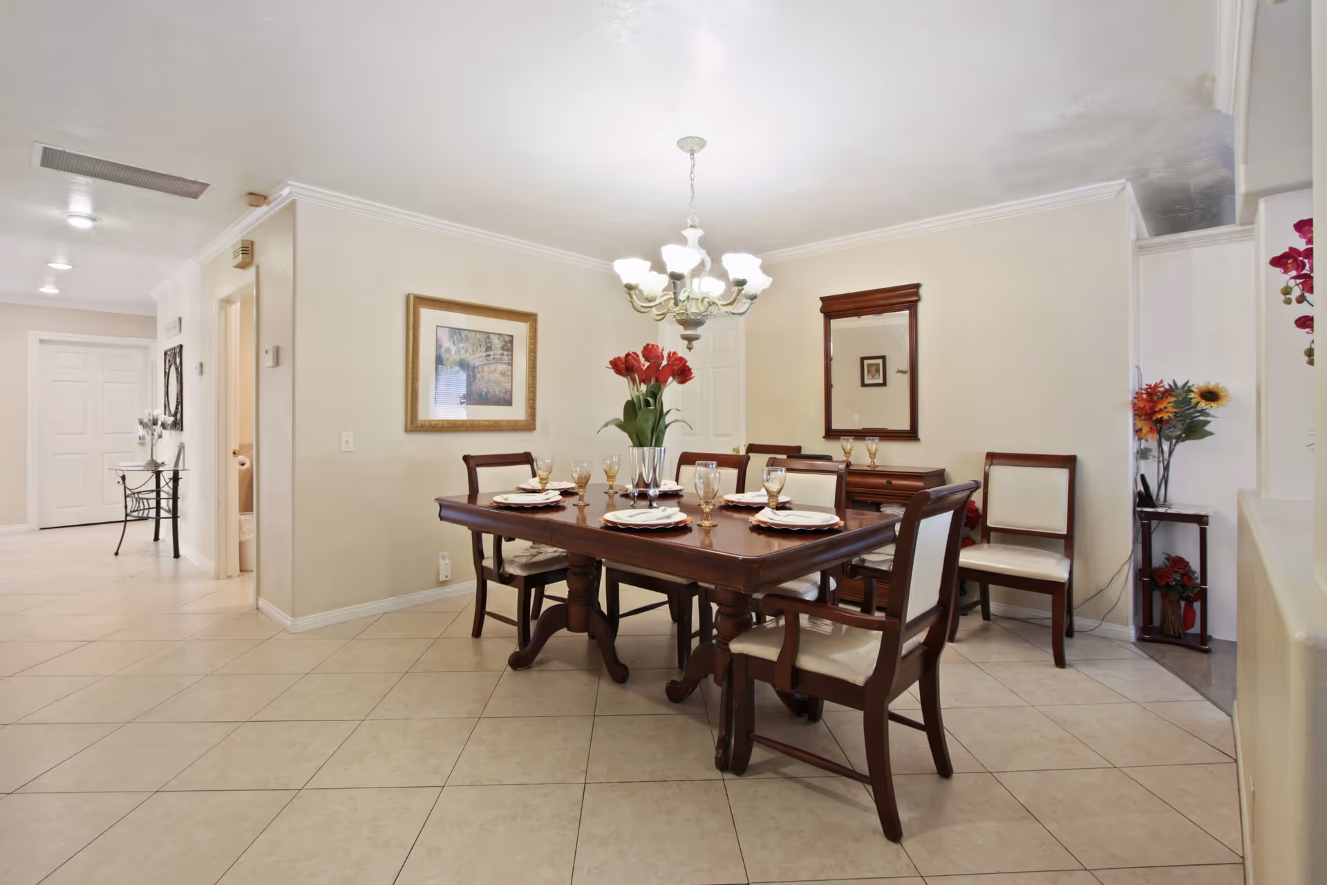 A dining room with a wooden dining table set for six people with plates, glasses, and a vase of red flowers in the center. The room has beige tiled floors, cream-colored walls, a chandelier hanging above the table, a framed picture on one wall, a mirror on another, and additional chairs and small tables with flower arrangements in the background.