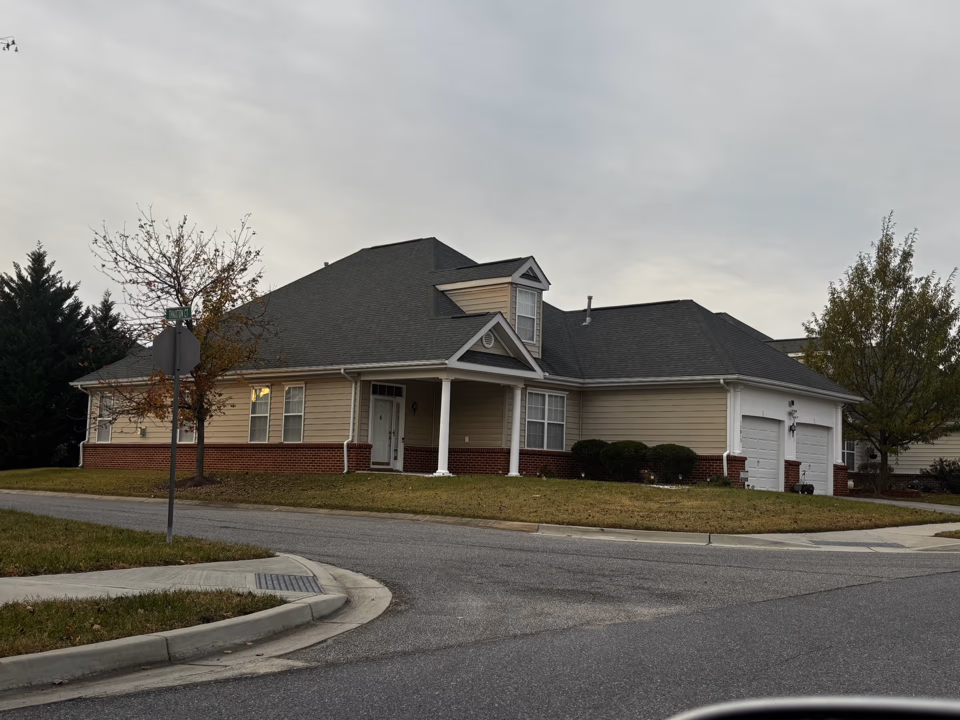 Exterior view of a single-story residential building with beige siding and a dark gray roof, featuring a small covered porch with white columns, several windows, a two-car garage, and a lawn with a few trees. The building is situated on a corner lot with a street sign visible.