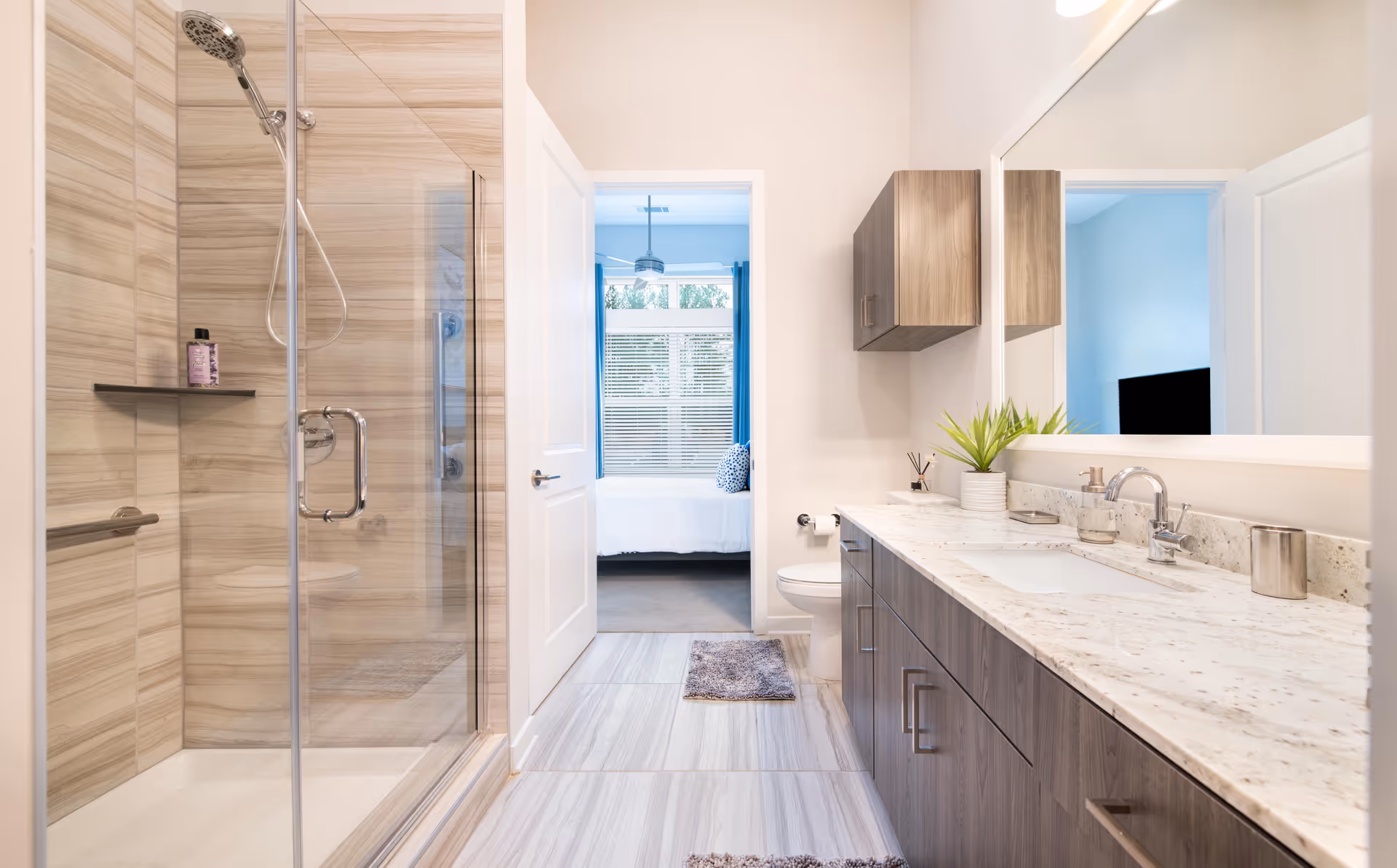 Modern bathroom with a glass-enclosed shower, long vanity with sink, and an open doorway leading to a bedroom.