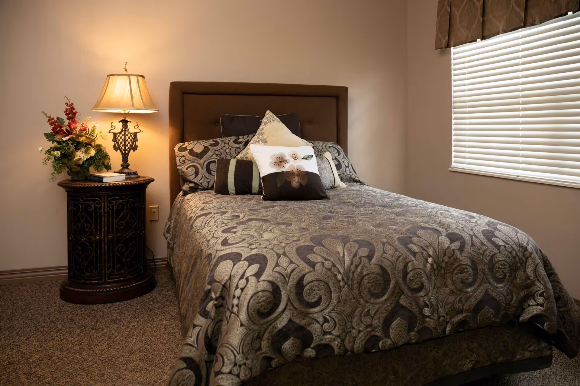 A neatly made bed with a patterned bedspread and multiple decorative pillows in a bedroom. To the left of the bed is a round wooden nightstand with an ornate lamp and a floral arrangement. A window with closed blinds is on the right wall.