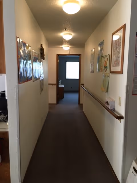 A long, narrow hallway with beige walls and carpeted floor. The hallway is lit by ceiling lights and has handrails on the right side. Various framed pictures and posters are hung on both walls. At the end of the hallway, there is an open doorway leading to a room with a window.