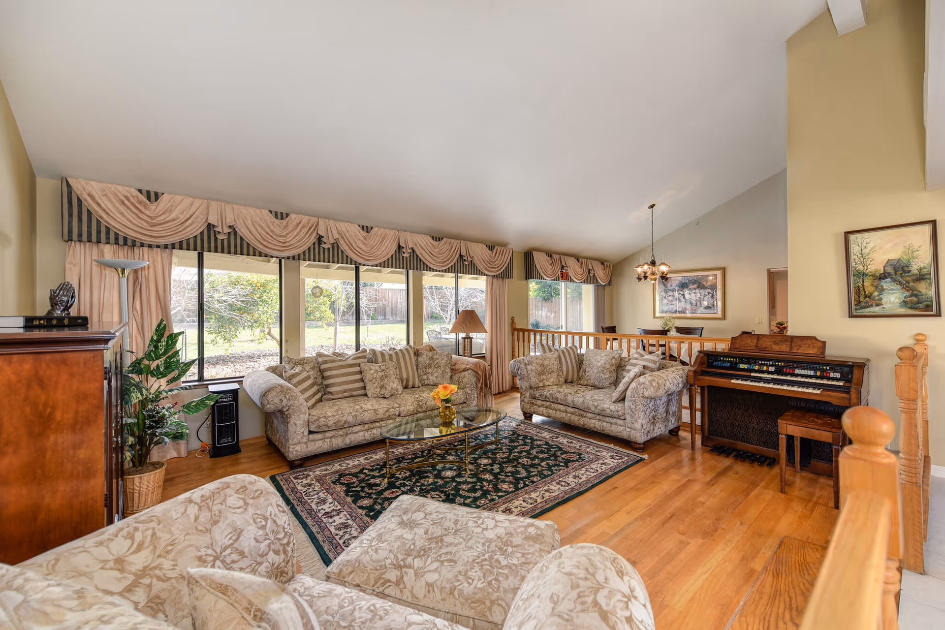 Bright living room with patterned sofas, a glass coffee table on an area rug, large windows with draped valances, and an organ against the wall.