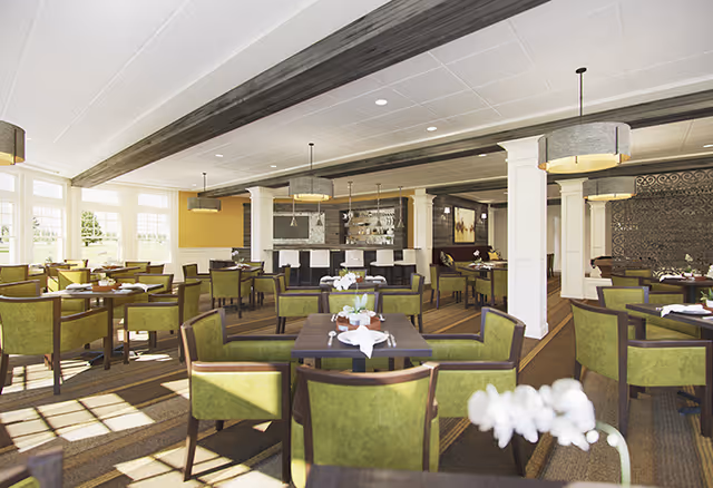 Bright, spacious dining room with multiple tables and green chairs arranged around a central bar and columns.