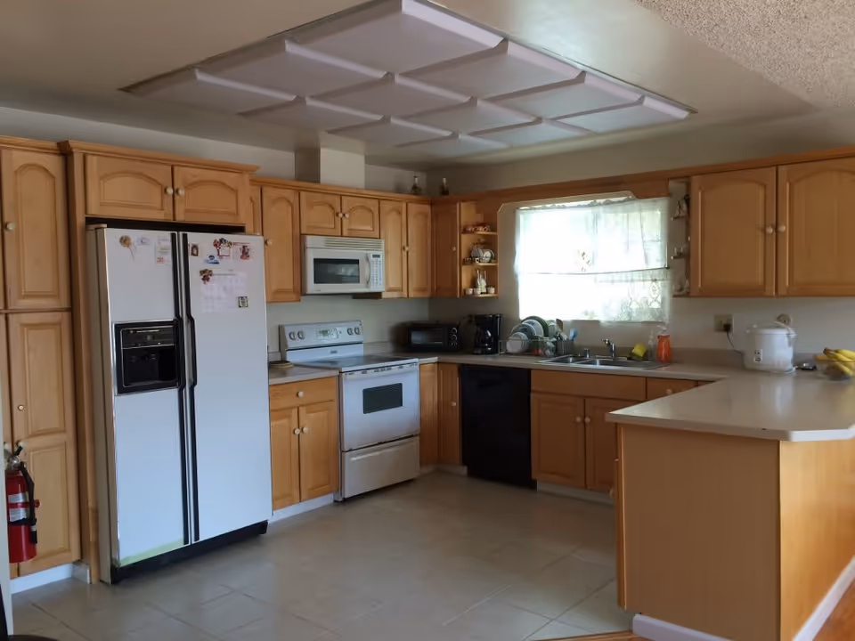 A kitchen with light wood cabinets, a white refrigerator with magnets, a white stove with an overhead microwave, a black dishwasher, a double sink under a window with lace curtains, and a countertop with a rice cooker and bananas.