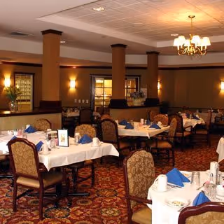 A dining room with multiple tables covered in white tablecloths, each set with blue napkins, cups, and silverware. The room features patterned carpet, upholstered chairs, warm lighting from wall sconces and a chandelier, and decorative columns.