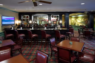 Interior view of a senior living facility lounge area with a bar counter, several high bar stools, and multiple tables with chairs. The room features a patterned carpet, a ceiling fan, a wall-mounted TV, decorative shelves behind the bar, and a large mural on one wall.