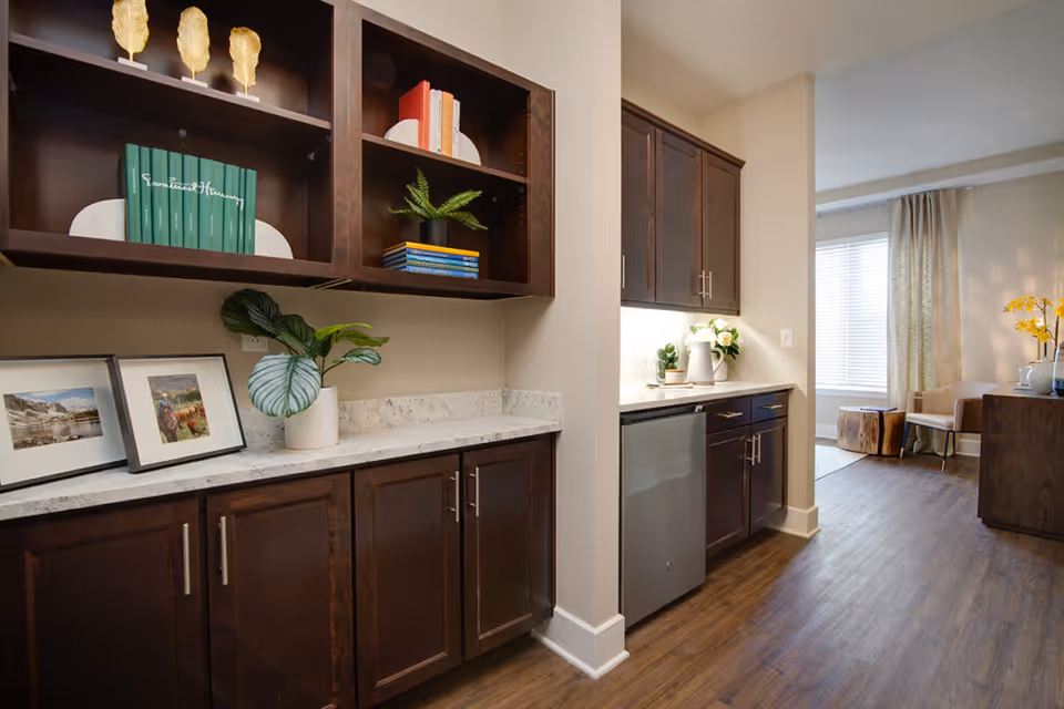Interior view of a senior living facility showing a hallway with dark wooden cabinets and white marble countertops. The cabinets have decorative items including framed pictures, books, plants, and golden leaf ornaments. The hallway leads to a living area with a window covered by curtains, a chair, and a wooden side table with a vase of yellow flowers.