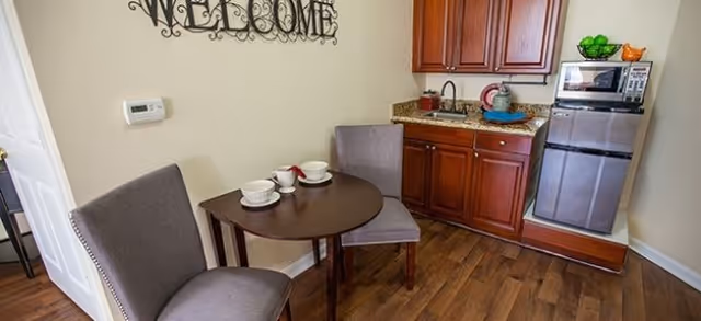 Small kitchenette and dining nook with two upholstered chairs, a round table, sink, wooden cabinets, and a mini-fridge beneath a "WELCOME" wall sign.