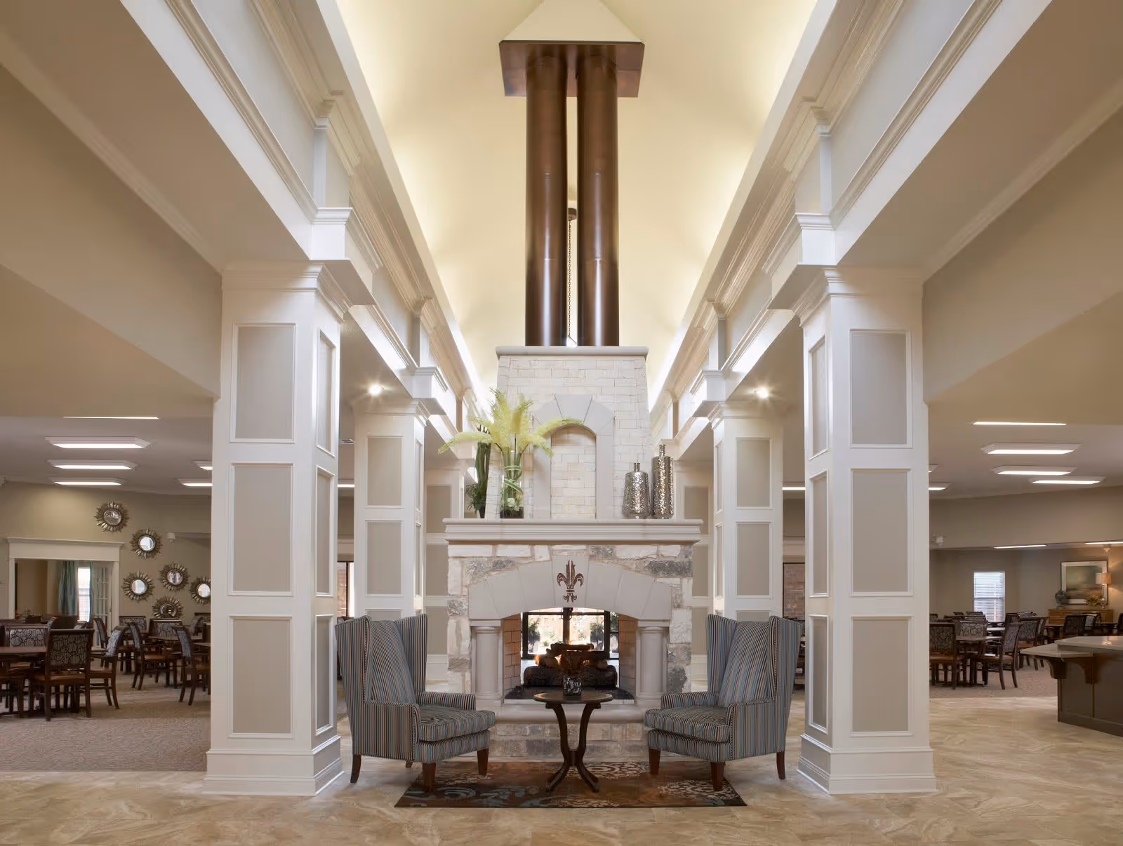 A spacious, well-lit common area with a large stone fireplace in the center, flanked by two striped armchairs and a small wooden table. The room features tall white columns, a high vaulted ceiling with recessed lighting, and multiple dining tables and chairs in the background.