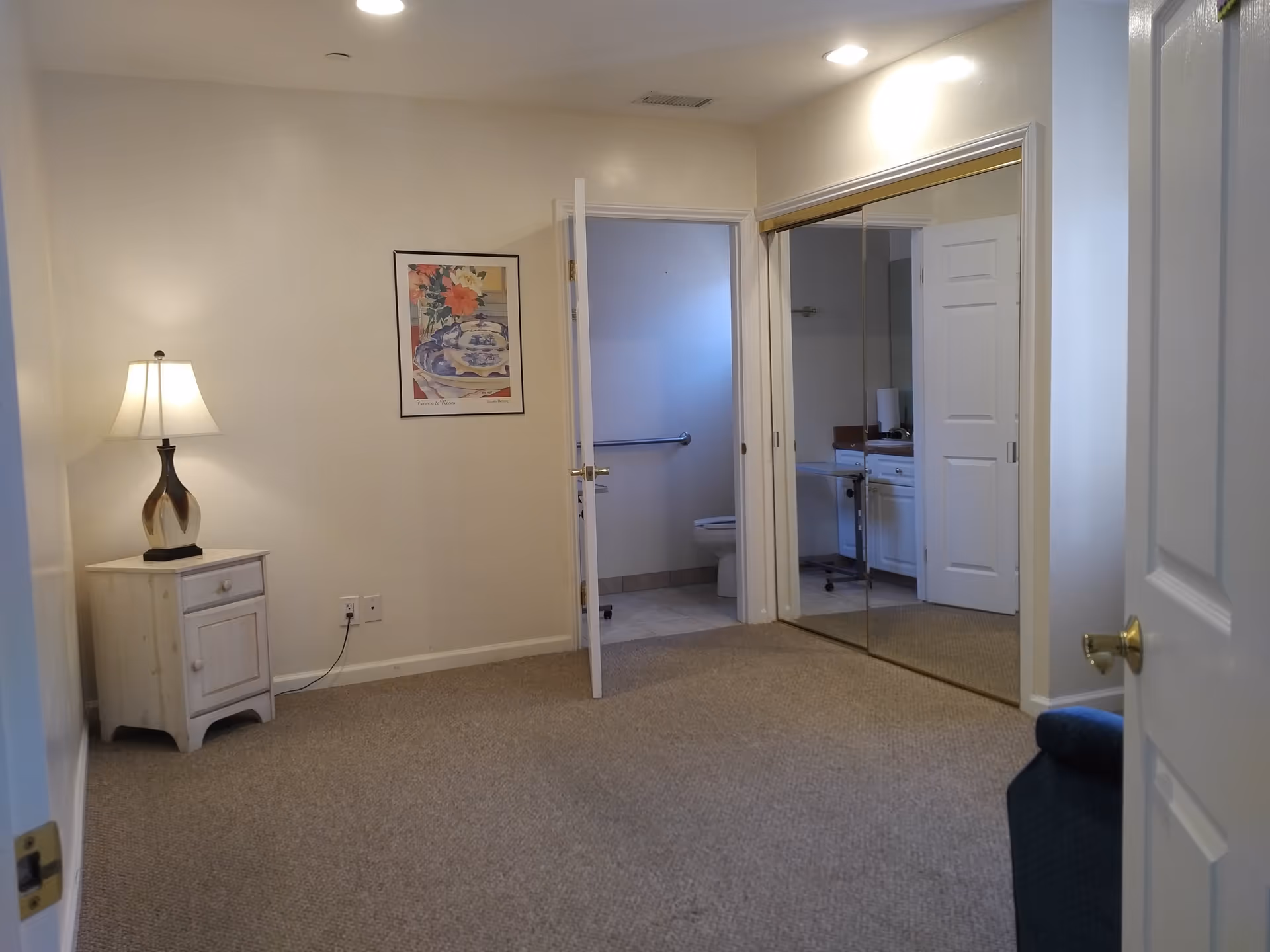 A carpeted room with beige walls featuring a small white nightstand with a lamp on it, a framed floral picture on the wall, a door leading to a bathroom with a visible toilet, and a large mirrored closet door reflecting part of the bathroom and room.