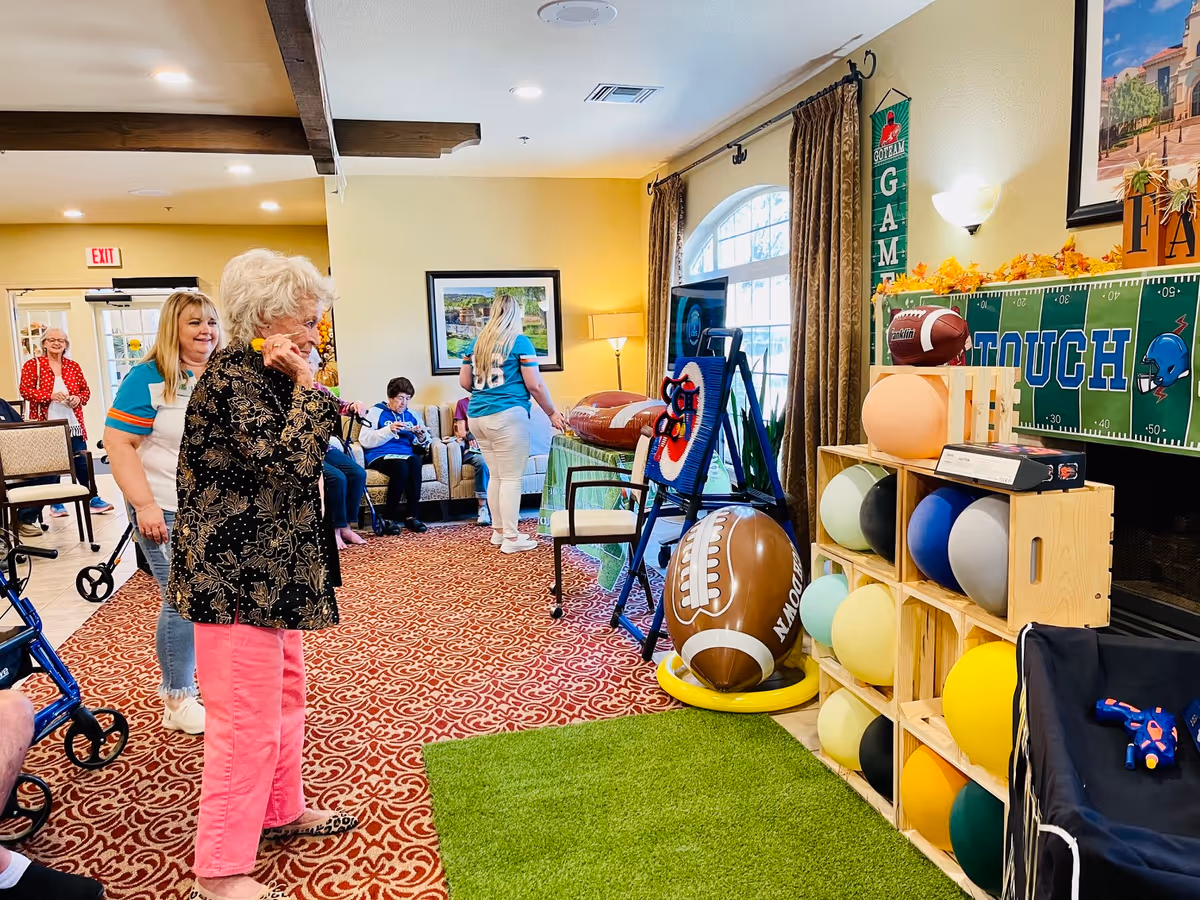 Residents and staff in a senior living common room playing games amid football-themed decorations and colorful exercise balls.