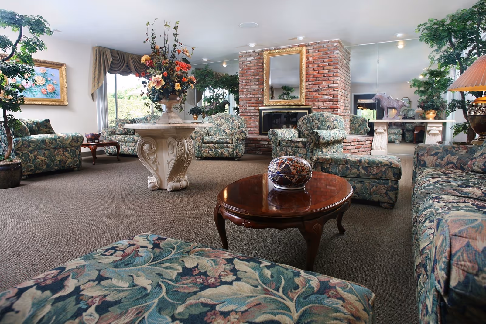 A spacious living room with floral patterned sofas and armchairs arranged around a wooden coffee table with a decorative ceramic piece on it. There is a brick fireplace with a gold-framed mirror above it, large indoor plants, a stone pedestal table with a large floral arrangement, and a painting on the wall. The room has large windows with drapes and mirrors reflecting the interior.