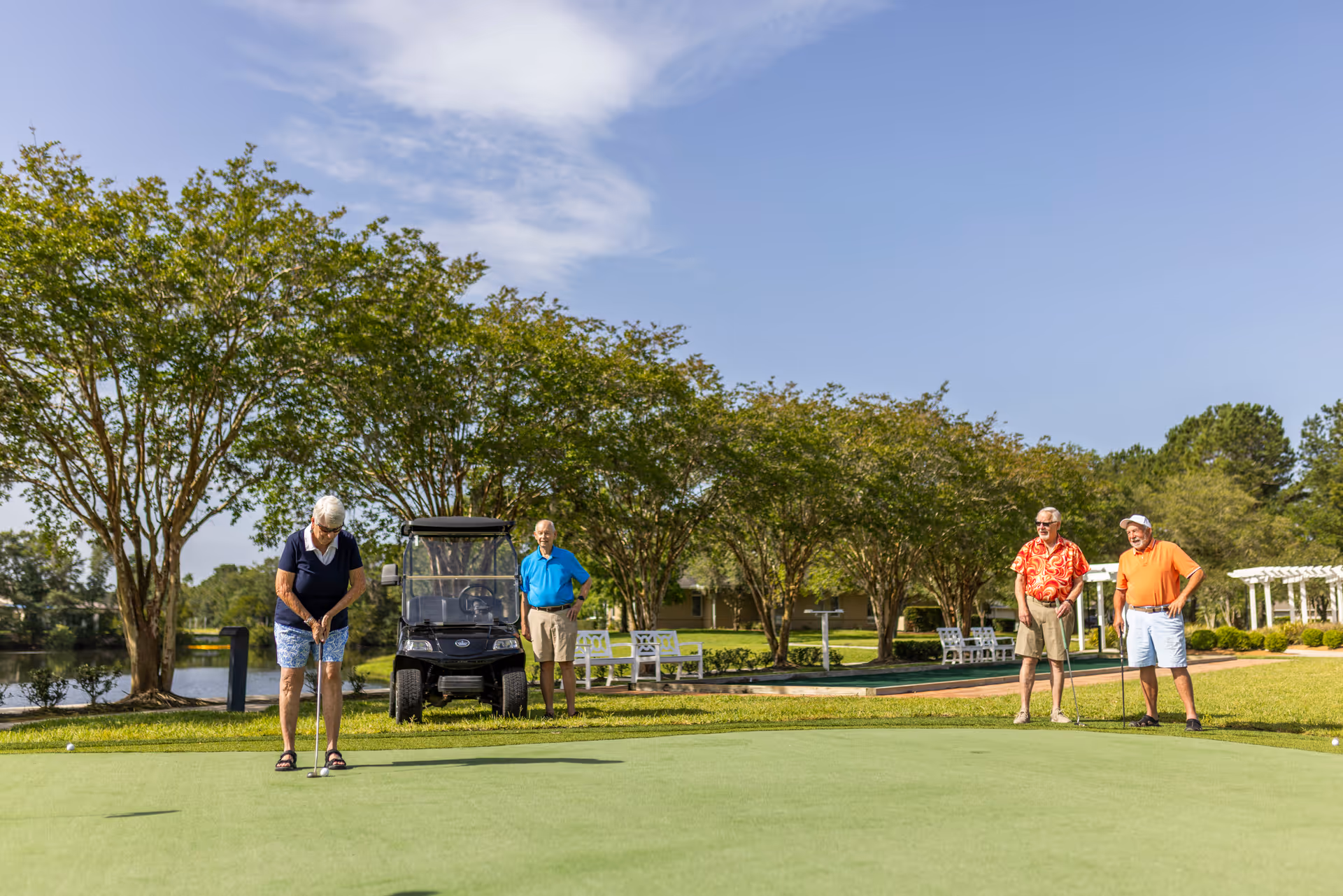 Four elderly people playing golf on a putting green at a senior living facility. One woman is putting while three men watch, with a golf cart and trees in the background under a clear blue sky.