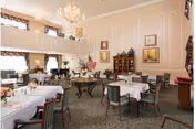 A spacious, elegant dining room with high ceilings, chandeliers, and large windows with curtains. The room is furnished with multiple tables covered with white tablecloths and set with dining ware. There are chairs around each table, and decorative paintings and furniture adorn the walls.