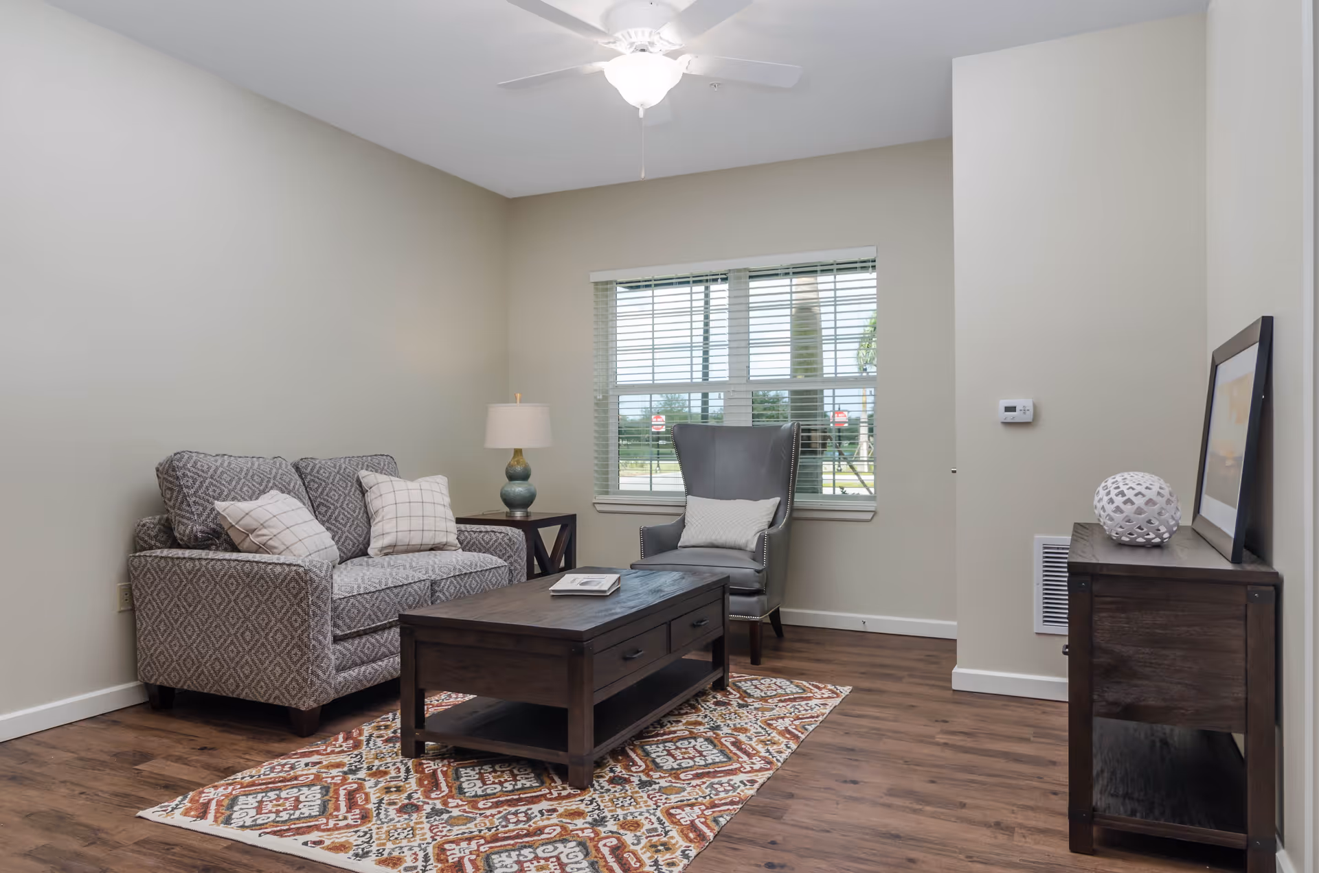 A cozy living room with a patterned loveseat and two pillows, a gray armchair with a small pillow, a wooden coffee table on a colorful patterned rug, a side table with a lamp, and a wooden console table with decorative items and a framed picture. A window with blinds lets in natural light, and a ceiling fan is mounted above.