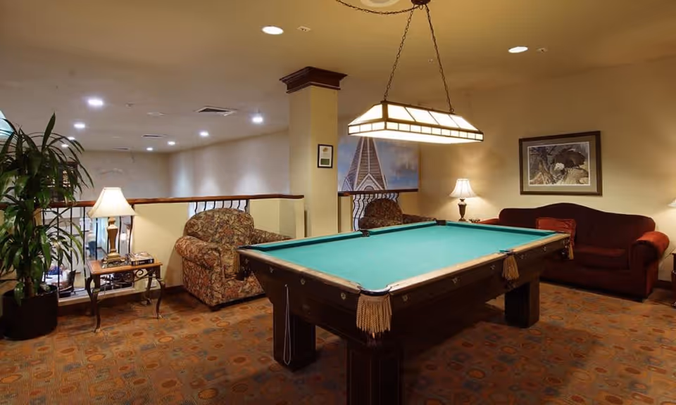 Cozy lounge with a pool table under a hanging light, surrounded by armchairs, a sofa, lamps, and a potted plant.