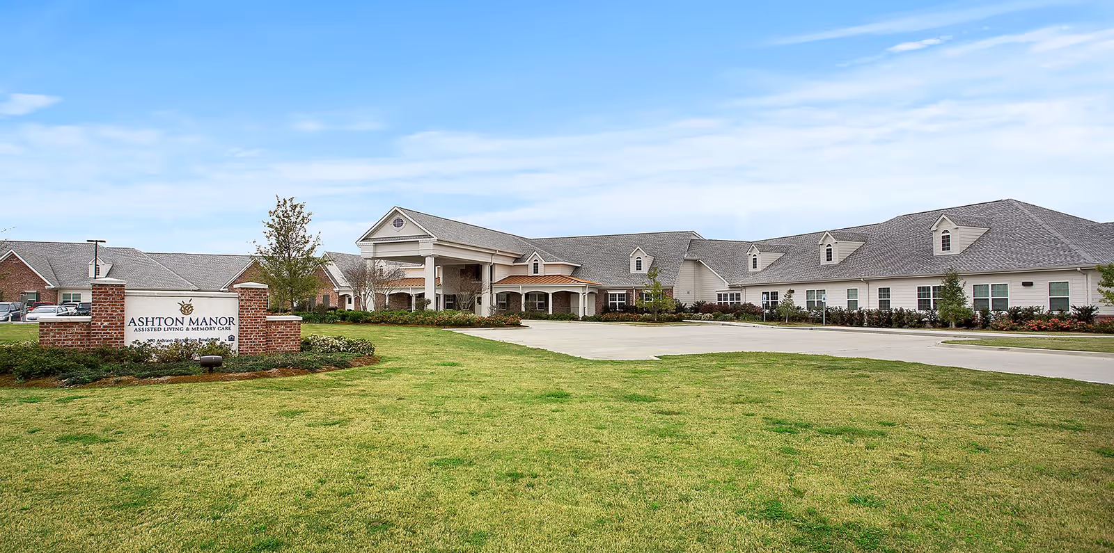 Exterior view of Ashton Manor assisted living and memory care facility showing a large building with a covered entrance, surrounded by a well-maintained lawn and landscaping under a partly cloudy sky.