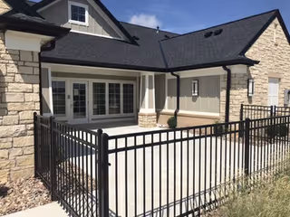 Exterior view of a senior care home building with stone and siding walls, a black shingled roof, and a fenced patio area under a clear blue sky.