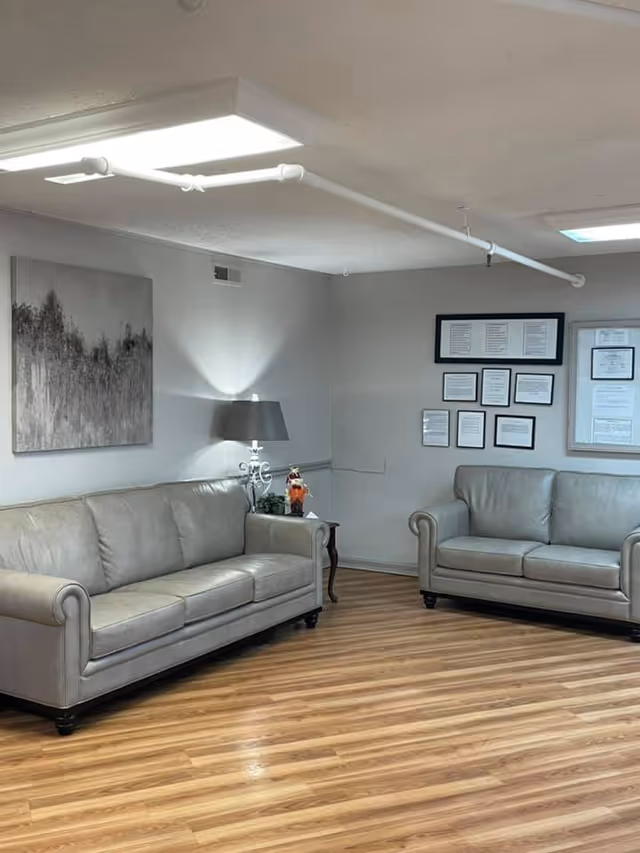 A lounge-style room with two gray leather sofas, a side table with a lamp and wall art on wood-look flooring.