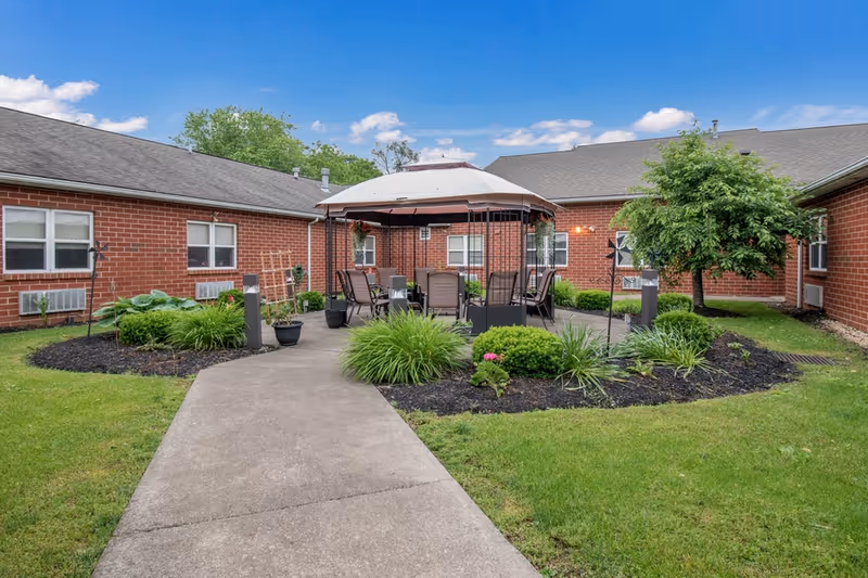 Outdoor courtyard area at Celebration Villa of Lewisburg featuring a concrete pathway leading to a gazebo with a canopy roof. The gazebo is surrounded by chairs and tables, with landscaped garden beds containing green shrubs and plants. The courtyard is enclosed by single-story red brick buildings under a blue sky with scattered clouds.