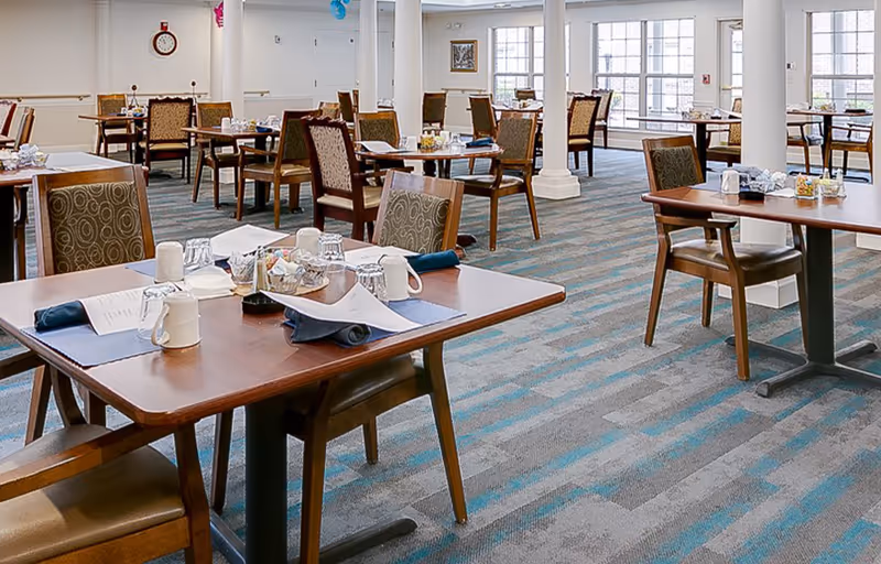 Bright senior living dining room with multiple tables and chairs set for meals and large windows.