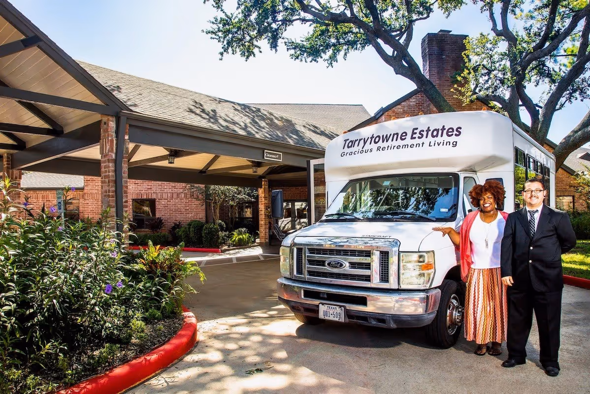 Two people standing next to a white shuttle bus with the sign 'Tarrytowne Estates Gracious Retirement Living' in front of a brick building with a covered driveway and surrounding greenery.