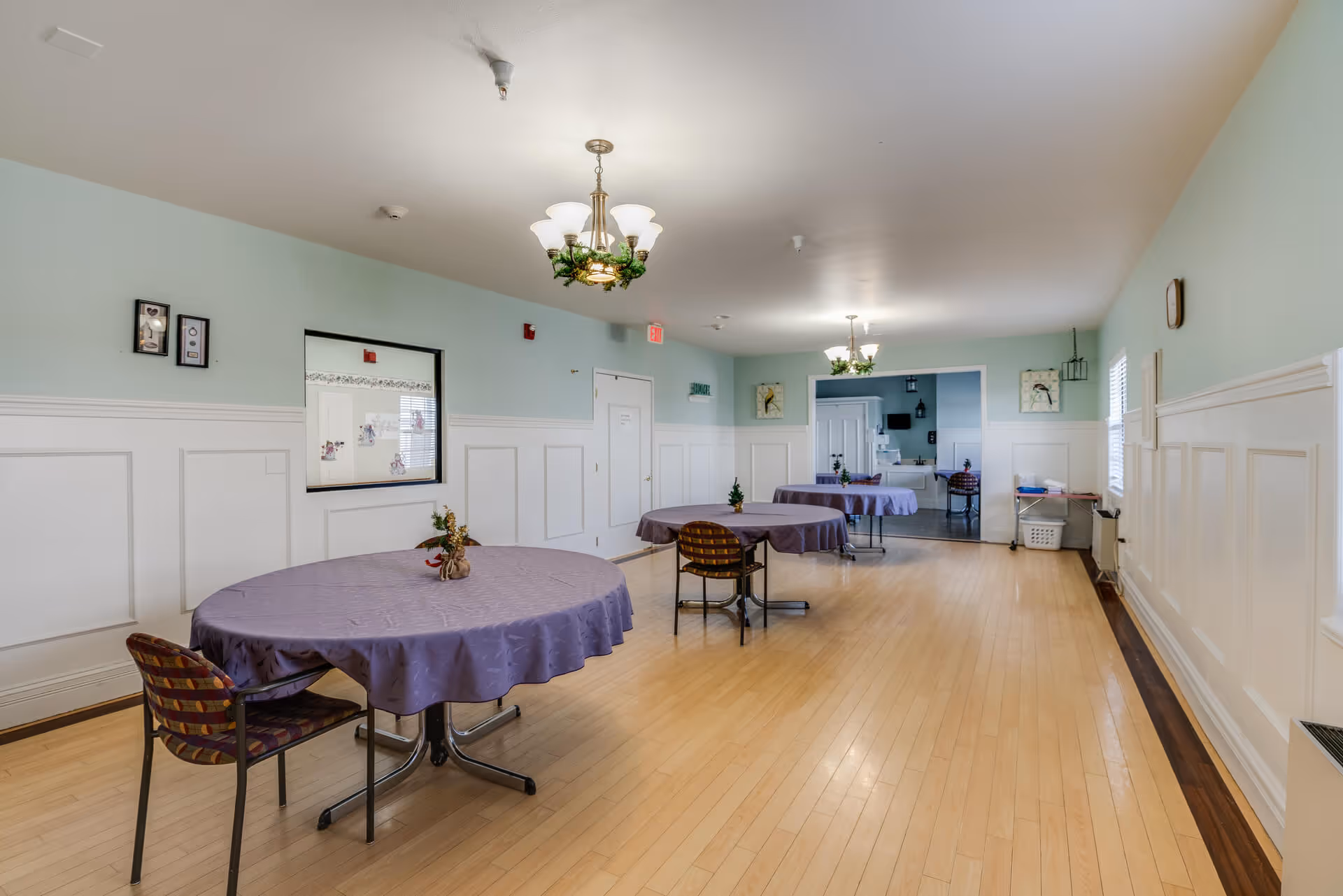 A spacious dining room with light blue walls and white wainscoting, featuring round tables covered with purple tablecloths and chairs around them. The room has wooden flooring, two chandeliers with greenery decorations, and a window opening to an adjacent room with a kitchen visible in the background.