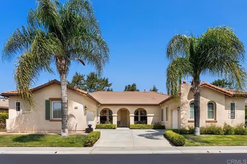 Front exterior of a single-story beige stucco building with a tiled roof, arched central entry, driveway and palm trees.