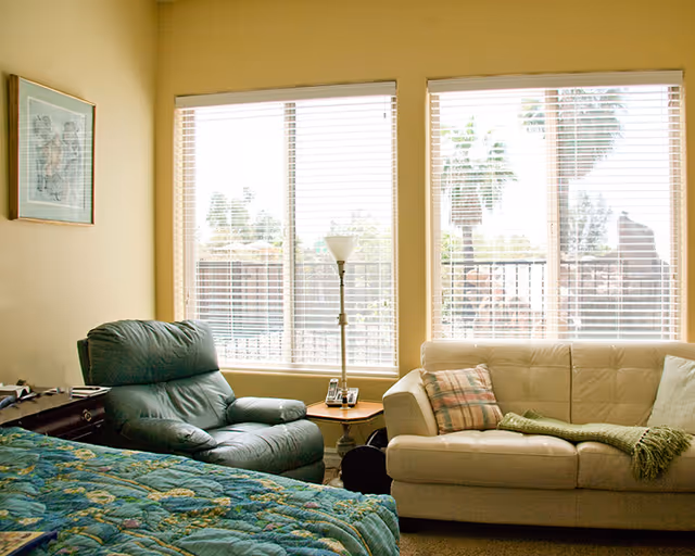 Sunlit room with a bed in the foreground, a green recliner, a cream sofa with pillows, and large windows with blinds.