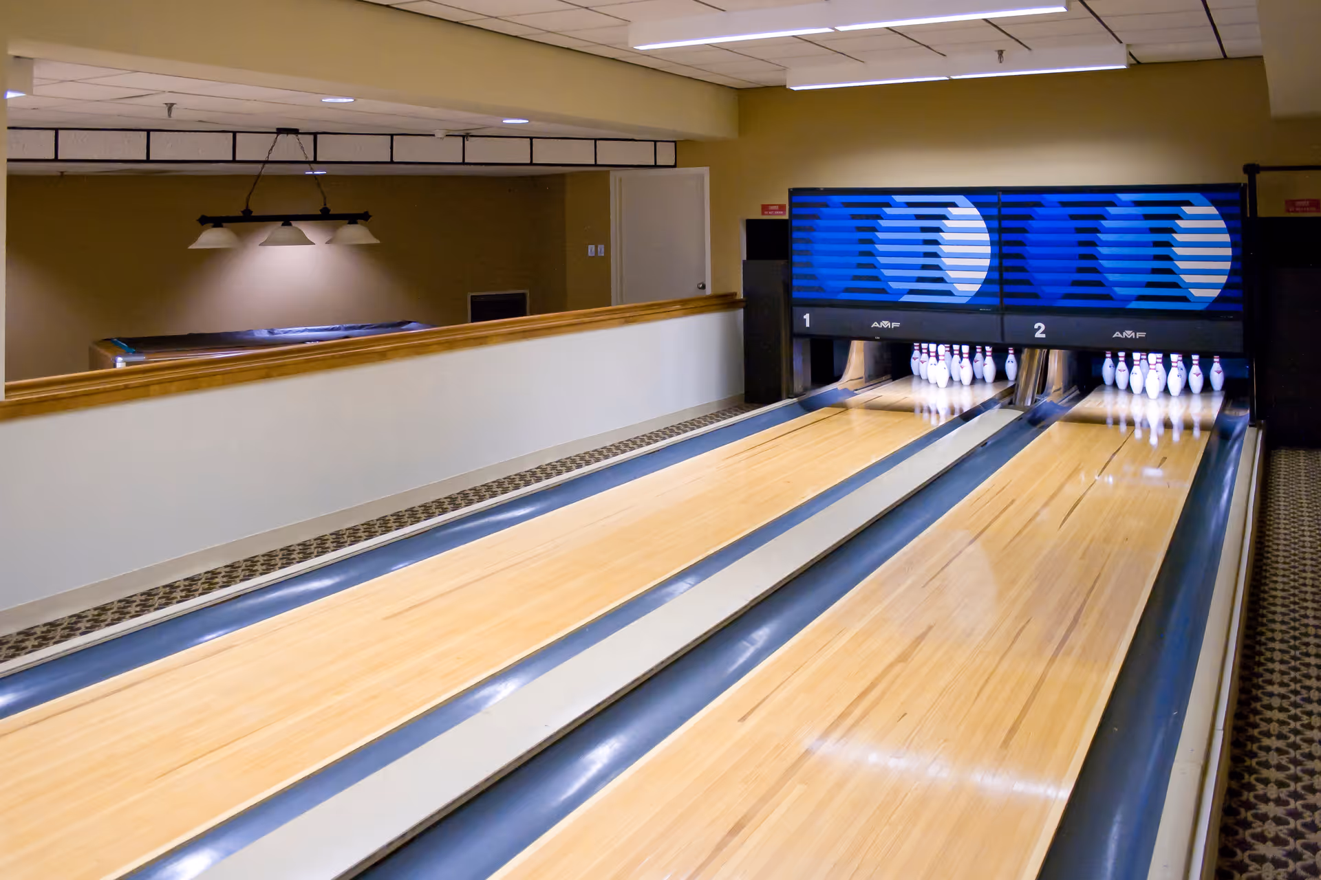 Indoor bowling alley with two lanes, pins set up at the end, and a pool table visible in the background under hanging lights.