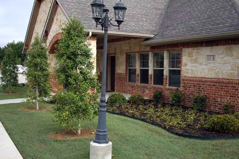 Exterior view of a brick and stone building with a sloped roof, several windows, a small garden with shrubs and trees, and a black street lamp on a grassy lawn.