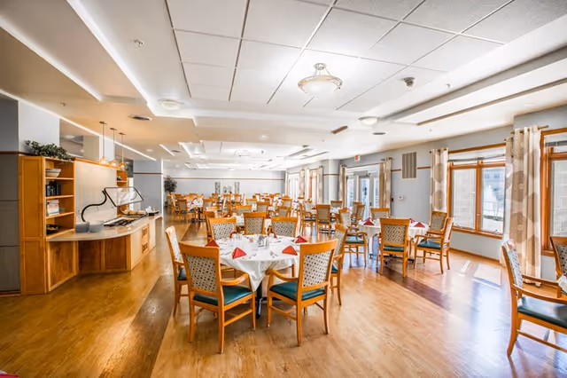A bright and spacious dining room with multiple round tables covered with white tablecloths and set with red napkins and silverware. Wooden chairs with patterned upholstery surround the tables. Large windows with patterned curtains allow natural light to fill the room. The floor is wooden, and there is a buffet area with a counter and shelves on the left side.