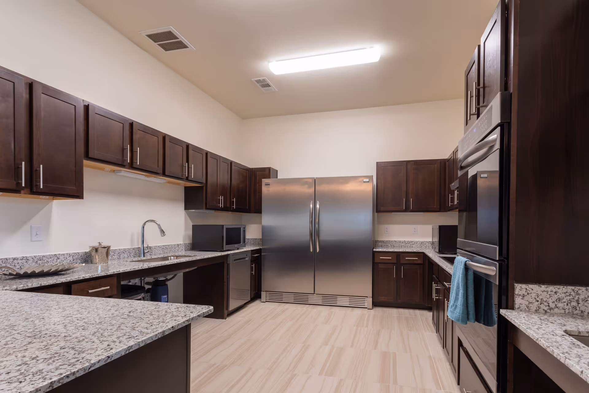 A modern kitchen with dark wood cabinets, granite countertops, a double-door stainless steel refrigerator, built-in microwave, dishwasher, and double wall ovens. The floor is tiled with light-colored tiles and the ceiling has a fluorescent light fixture.