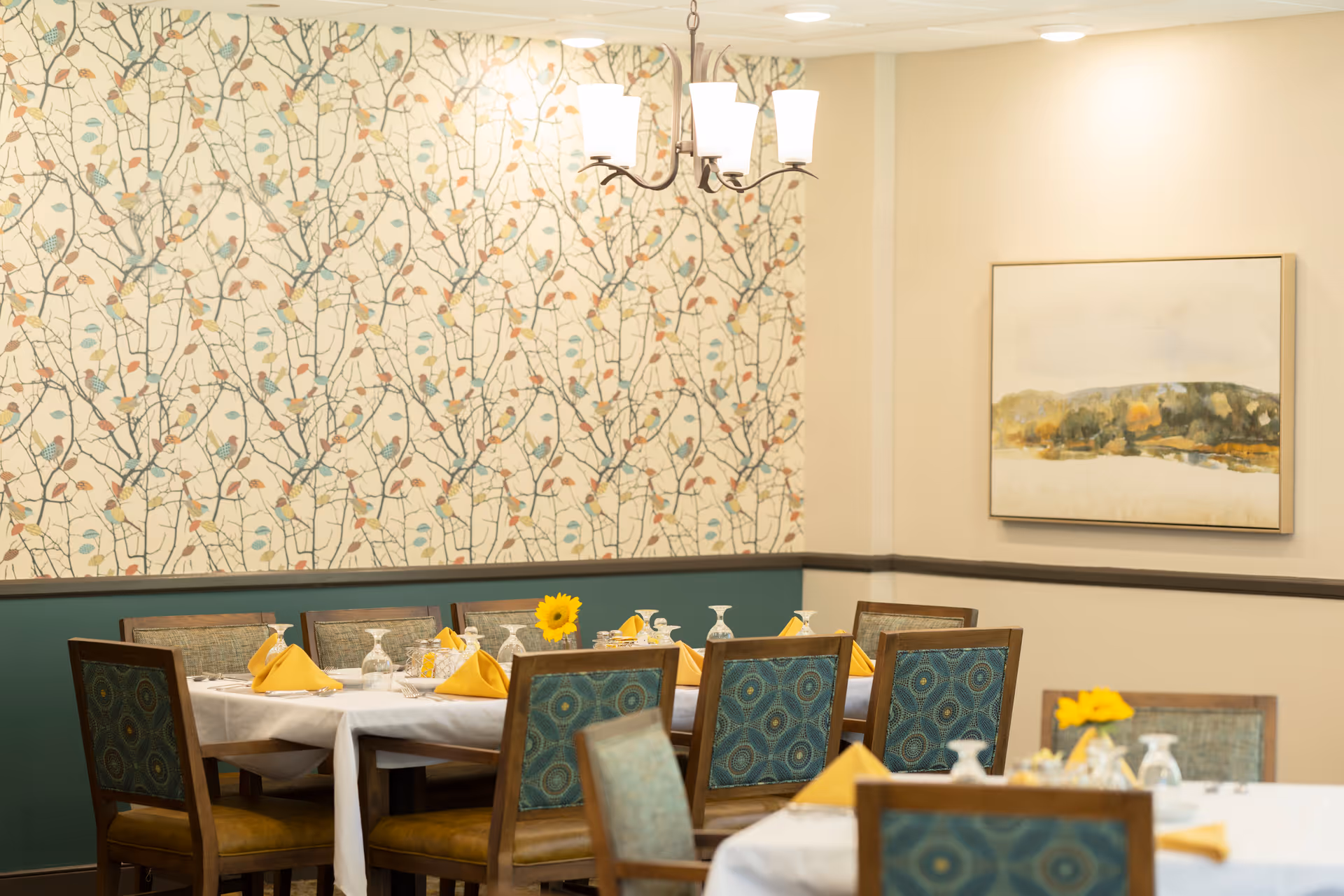 A dining room with a table set for a meal, featuring yellow napkins, glassware, and a small vase with a yellow flower. The room has patterned wallpaper with a branch and leaf design, a chandelier with three lights, and a framed landscape painting on the wall.