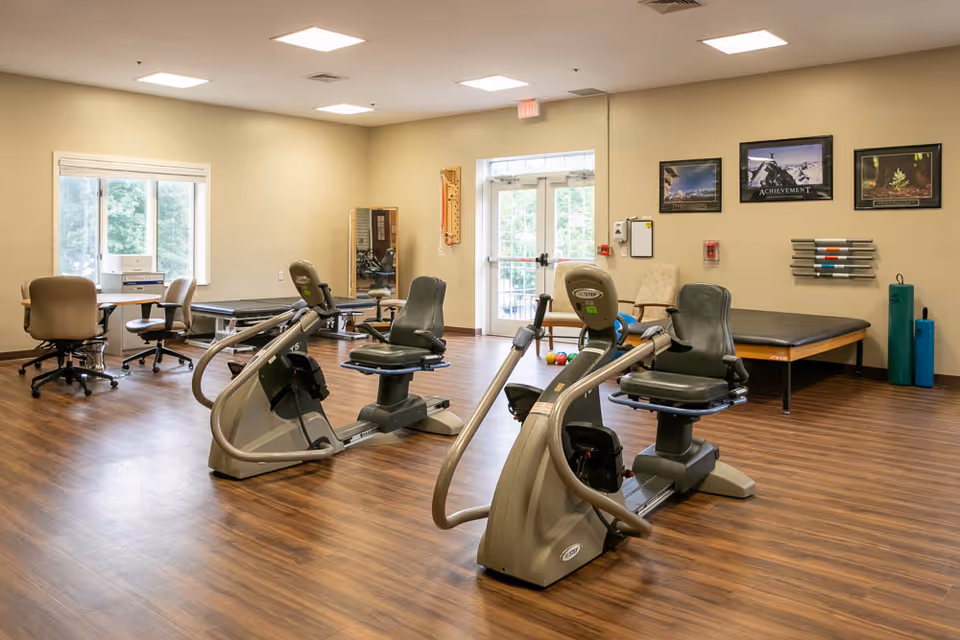 A spacious rehabilitation room with two recumbent exercise bikes in the center, a therapy table against the wall, a desk with chairs near a window, and various exercise equipment and motivational posters on the walls.