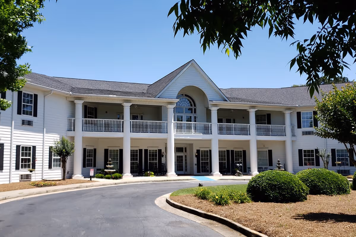 Front exterior view of a two-story senior living facility building with white siding, large white columns, black shutters, and a curved driveway leading to the entrance. There are manicured bushes and trees in the landscaped area in front of the building under a clear blue sky.
