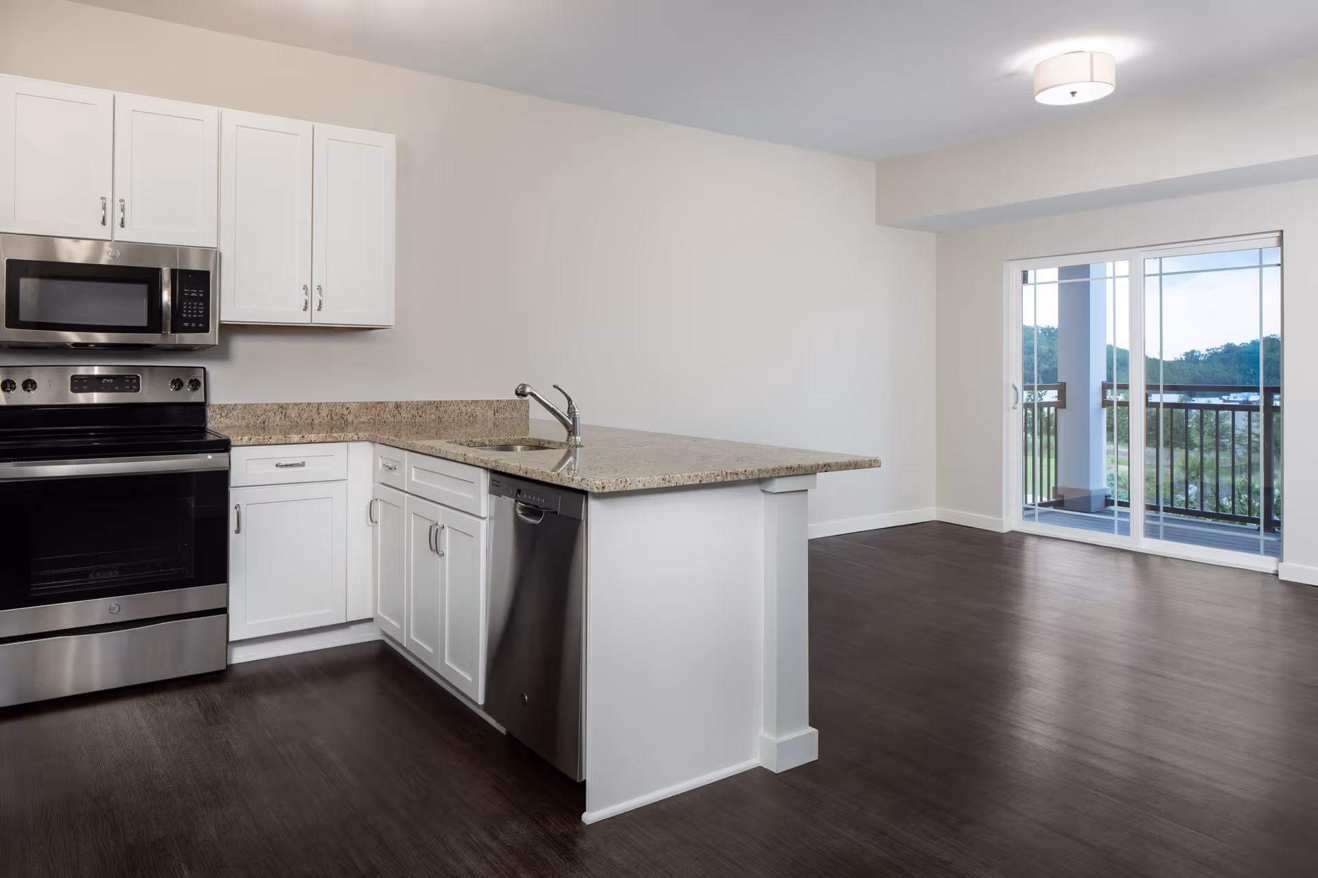 Modern kitchen with white cabinets, stainless steel appliances including a microwave, oven, and dishwasher, granite countertops, and a sink. The kitchen opens to a living area with dark wood flooring and a sliding glass door leading to a balcony with a view of greenery and hills.